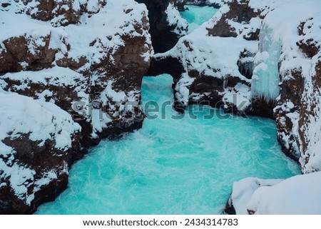 Winter stream with turquoise water flowing through snow-covered rocks and under a natural bridge. Location: Hraunfossar, Iceland.