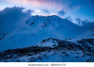 A winter summit tearing through cloud crowned with pink dawn light above a frost-rimed foreground — a hero winter mountain shot.