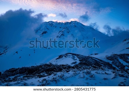 A winter summit tearing through cloud crowned with pink dawn light above a frost-rimed foreground — a hero winter mountain shot.