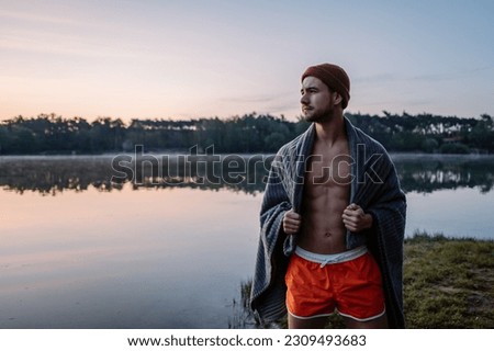 Winter swimming. Serious guy with towel at his shoulders walking at the forest near the water and preparing for the swimming in the cold winter lake at morning