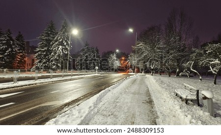 In winter, there is snow on the streets and trees. The sidewalks and roadways have been cleared by public utilities. Darkness fell. Cars with headlights are driving. Lights are burning on poles