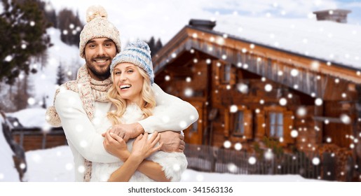 Winter, Vacation, Couple, Christmas And People Concept - Smiling Man And Woman In Hats And Scarf Hugging Over Wooden Country House And Snow Background