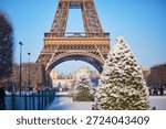 A winter view of the Eiffel Tower in Paris with symmetrical rows of snow-covered trees in the foreground. Classic seasonal scene with clear blue sky, perfect for Christmas and New Year themes.