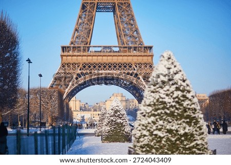 A winter view of the Eiffel Tower in Paris with symmetrical rows of snow-covered trees in the foreground. Classic seasonal scene with clear blue sky, perfect for Christmas and New Year themes.