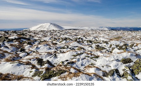 Winter view in the mountains. In the foreground are stones in the snow. A snow-capped mountains in the distance. Clear blue sky.