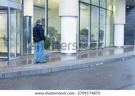 Winter walk beside large windows of modern structure as man secures outerwear in falling snow on reflective sidewalk. High quality photo