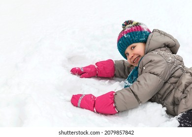 Winter Walking Activity. Girl Lying In Snow And Playing. Child Kid In Winter Suit, Knitted Hat And Scarf And Waterproof Gloves. Looking In Camera