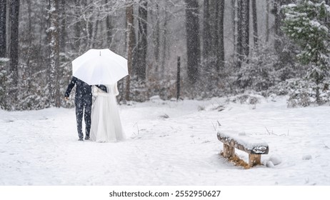 Winter wedding scene of a couple walking through a snowy forest under an umbrella, surrounded by snow-laden trees - Powered by Shutterstock