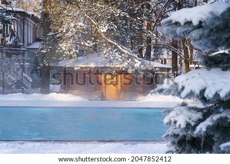 Winter wellness spa resort with hot outdoor swimming pool with thermal water streaming in morning frozen air surrounded with trees in snow near beauty salon or hotel. Body care and relaxation concept