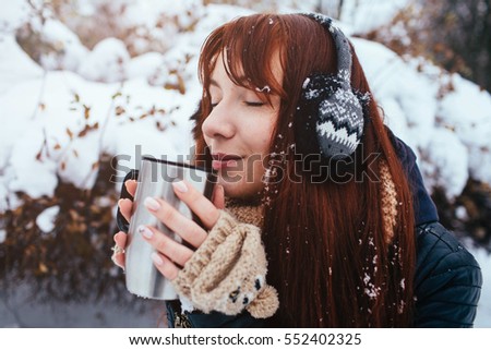 Winter. Woman with red hair wearing ear muffs. Girl drinking hot tea or coffee  iron insulated cup