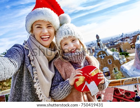 Winter wonderland in Barcelona at Christmas. happy trendy mother and daughter travellers with a little Christmas tree and present box at Guell Park in Barcelona, Spain taking selfie
