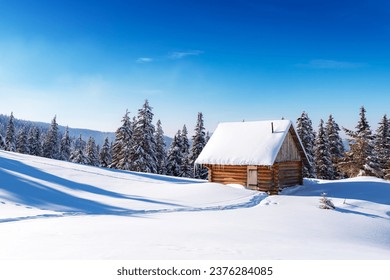 A wintery scene with a mountain meadow, snow-covered pine trees, and a rustic wooden cottage. Clear blue sky on background - Powered by Shutterstock