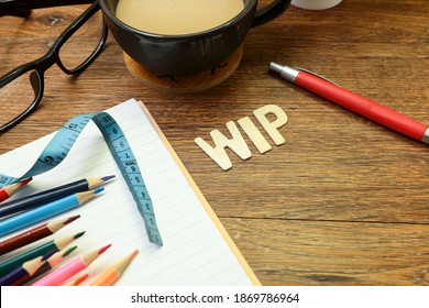 WIP, Work In Progress concept.  Close view of block letters on WIP on the wooden table with a pen, notebook, group of pencils, a cup of coffee, eyeglasses and measuring tape 