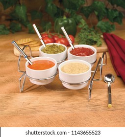 Wire Condiment Tray Including Ketchup, Mustard, Relish, French Dressing, With Spoons On Butcher Block With Tomatoes And Peppers In Background.