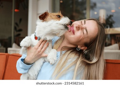 Wire haired jack russell terrier licking a cheek of his joyful woman outside of the coffee shop. Young woman and her dog playing outdoors. Copy space, background. - Powered by Shutterstock