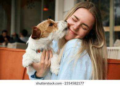 Wire haired jack russell terrier licking a cheek of his joyful woman outside of the coffee shop. Young woman and her dog playing outdoors. Copy space, background. - Powered by Shutterstock