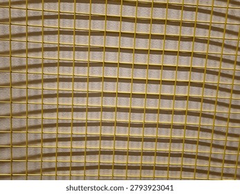 Wire mesh casting shadows on a yellow wall background, creating geometric patterns and textured surfaces suitable for abstract and design concepts.