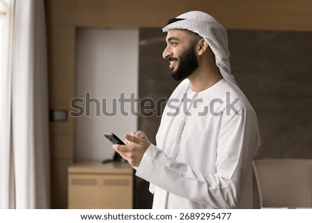 Wireless connection. Smiling young Arabic man in traditional Islamic white robe student employee relax in office with phone text message in chat play web game communicate online using wifi. Copy space