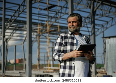 Wireless device, holding digital tablet in hands. Senior worker is near the unfinished greenhouse, working. - Powered by Shutterstock