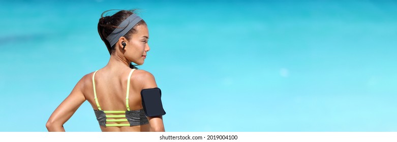 Wireless earphones and mobile phone music app for runner athlete running Asian woman listening to music podcast with earbuds wearing armband sleeve on beach run workout. Healthy fitness girl banner. - Shutterstock ID 2019004100