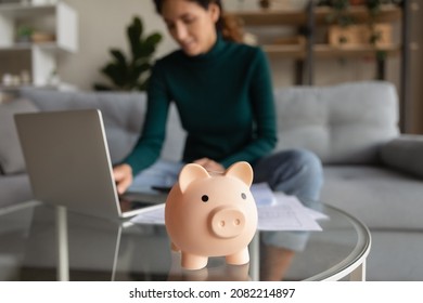 Wise economy. Blurred cropped shot of young woman managing household budget at home office planning family finances paying utility bills using app on laptop. Focus on funny pink piggybank toy on table