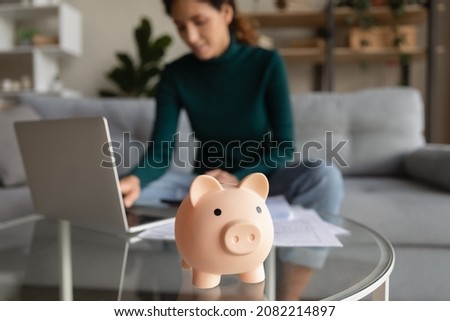 Wise economy. Blurred cropped shot of young woman managing household budget at home office planning family finances paying utility bills using app on laptop. Focus on funny pink piggybank toy on table
