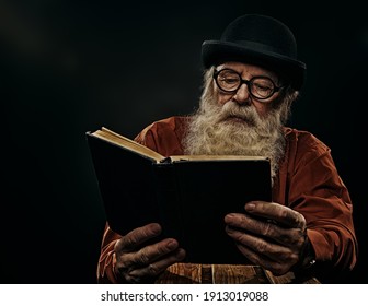 A wise old man with a long gray beard in a bowler hat and glasses is reading an old book. Black background. 