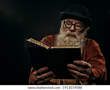 A wise old man with a long gray beard in a bowler hat and glasses is reading an old book. Black background. 