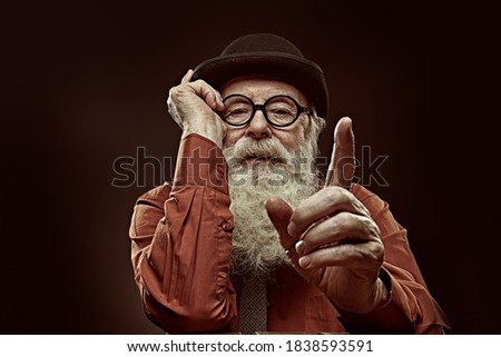 A wise old man with a long white beard in a bowler hat and glasses gives advice and shows his index finger. Black background. 
