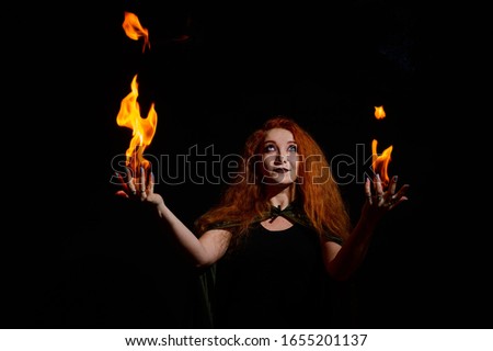 A witch with long curly hair holds a magic fire. Tongues of flame on the palms of a red-haired woman.