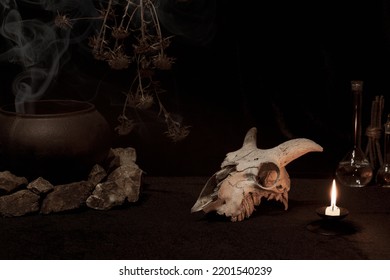 Witchcraft, Pagan Altar. Goats Skull, Burning Candle, Dry Herbs, Iron Cauldron, Smoking Incense On Black Background, Low Key, Selective Focus. Samhain, Magic Rite, Voodooo, Occultism, Alchemy Concept