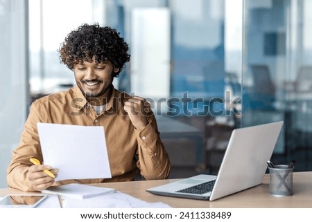 Witness the captivating essence of a vibrant Indian man with curly hair, fully immersed in his work as he sits at a table, surrounded by scattered papers, alongside his trusty laptop.