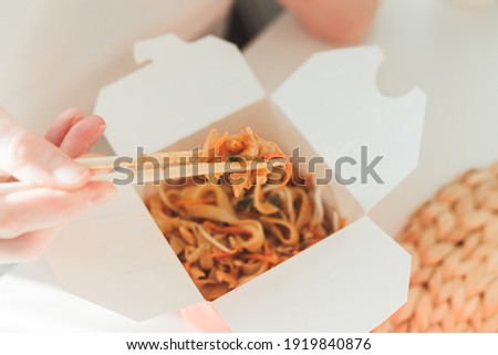 Wok noodles in takeaway box. Woman eating with chopsticks, close up view on female hands. Chinese traditional food with vegetables and seafood.