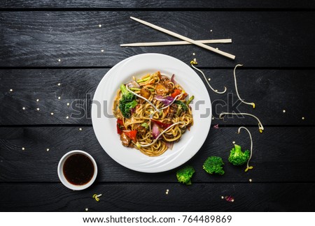 Wok. Udon stir fry noodles with chicken and vegetables in a white plate on black wooden background. With chopsticks and sauce