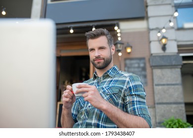 Woking Remotely. Bearded Handsome Male Ejoying Coffee, Looking At His Laptop In Open Air Cafe