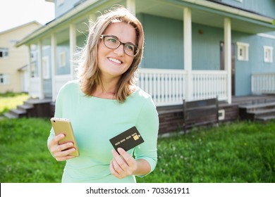 Woman of 30 years old near the house holds a phone and a credit card. Idea for app for renting or buying a house, an apartment. 