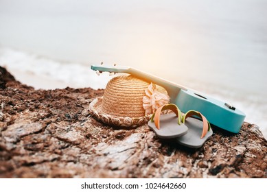 Woman accessories in vacation at beach. Ukulele and sandals and hat on rock beside sea. Tropical and season concept. Summer and holiday theme. Relaxation and Nature theme.