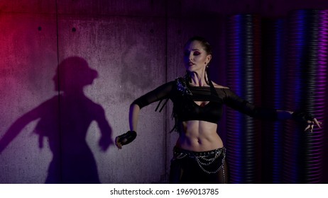 A woman is actively dancing in the studio against the background of the wall from which her shadow is reflected, she is dressed in a suit with an open stomach and is illuminated by light.
