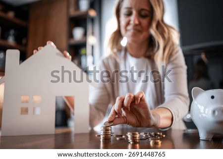 Woman adding coins to a stack, representing financial growth and investment toward buying real estate, with a house model and piggy bank symbolizing homeownership and future planning