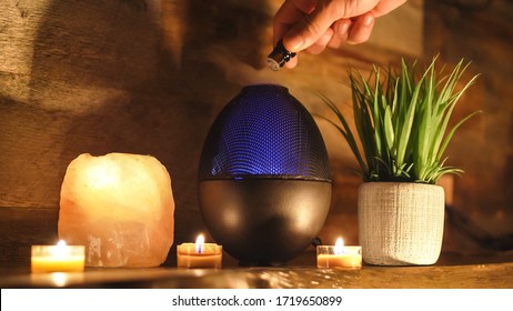 Woman adding essential oil to electric diffuser lamp, candles on wooden table in room. Aromatherapy.
