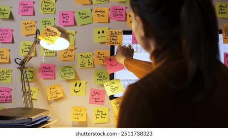 Woman adds a colorful smiley face paper to a computer screen surrounded by many other colorful sticky notes, working late in the evening - Powered by Shutterstock