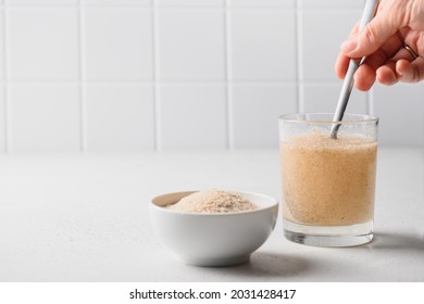 Woman adds spoon psyllium fiber and mix up in glass of water on a kitchen white background. Superfood for healthy intestines and gluten free diet.