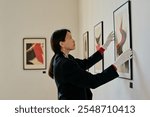 Woman adjusting framed artwork on gallery wall wearing gloves. Modern abstract paintings hanging alongside for exhibition