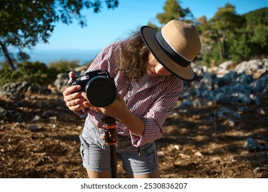 A woman adjusting her camera on a tripod while enjoying a photography session outdoors, surrounded by natural scenery and sunlight. She wears a straw hat and a checkered shirt. - Powered by Shutterstock