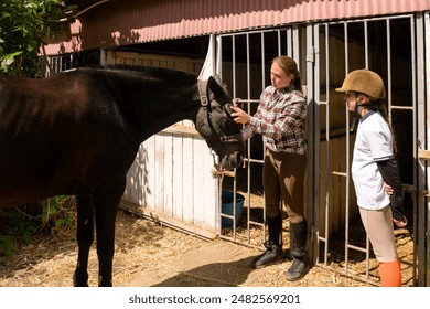 Woman adjusting horse's bridle as child girl observes at a stable. - Powered by Shutterstock