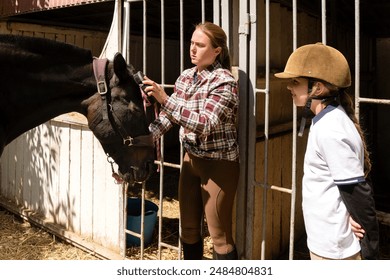 Woman adjusting horse's bridle as child girl observes at a stable. - Powered by Shutterstock