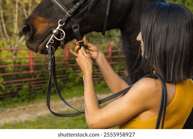 Woman adjusting a horse's bridle strap outdoors. Lifestyle. Horse riding. - Powered by Shutterstock