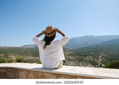 Woman admiring scenic countryside view, sitting on a stone ledge with mountains in the background on a sunny day - Powered by Shutterstock