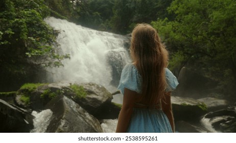 Woman admiring scenic waterfall surrounded by lush greenery and rocks, back view, taken in tranquil forest. Nature and Travel. - Powered by Shutterstock