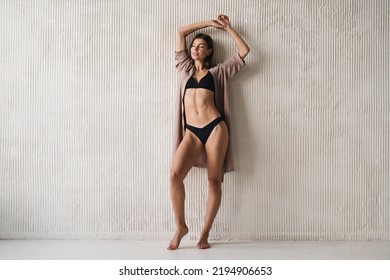 Woman Advertising New Lingerie Collection Standing Near Wall With Closed Eyes And Hands Raised Up, Dressed In Cotton Dressing Gown And Black Bikini. Home Outfit Clothing